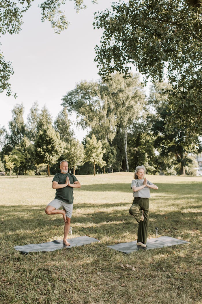 Elderly couple practicing yoga poses on mats in a sunny park setting.