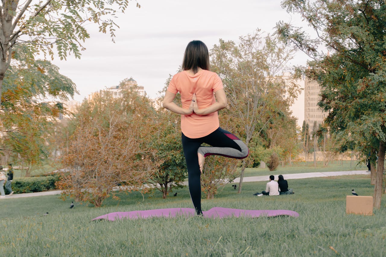 Adult woman performing yoga on a mat in a park, surrounded by greenery and trees.